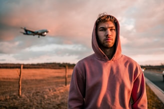 Person wearing a cozy streetwear hoodie with airplane motifs, standing near an airport runway at sunset
