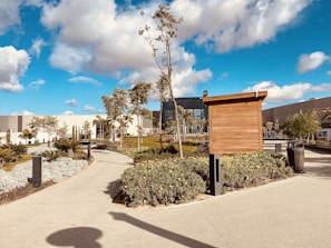 A landscaped outdoor area with a pathway winding through well-maintained gardens. The scene includes various types of plants and shrubs, along with a few tall trees. A wooden structure stands prominently near the path. In the background, a modern building with glass windows and a geometric design is visible. The sky is bright with scattered clouds, suggesting a sunny day.