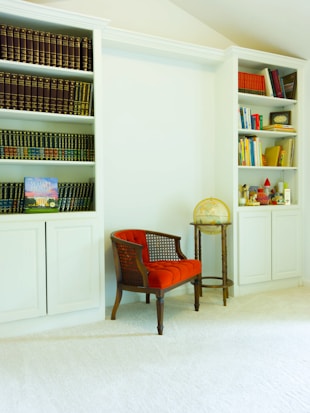 A bright and cozy reading nook features two built-in bookshelves filled with a variety of books and decorative items. The left shelf contains numerous leather-bound books, while the right shelf holds colorful books and various trinkets. A small wooden chair with a vibrant red cushion is placed in front of the shelves, next to a side table that holds a small globe.