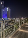 A modern cityscape at night featuring a tall building illuminated with blue and white lights, with the words 'Grand Fiesta Americana' prominently displayed. The foreground shows an elevated walkway with sleek, white railings and embedded ground lights.