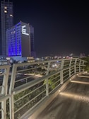 A modern cityscape at night featuring a tall building illuminated with blue and white lights, with the words 'Grand Fiesta Americana' prominently displayed. The foreground shows an elevated walkway with sleek, white railings and embedded ground lights.