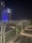 A modern cityscape at night featuring a tall building illuminated with blue and white lights, with the words 'Grand Fiesta Americana' prominently displayed. The foreground shows an elevated walkway with sleek, white railings and embedded ground lights.
