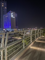 A modern cityscape at night featuring a tall building illuminated with blue and white lights, with the words 'Grand Fiesta Americana' prominently displayed. The foreground shows an elevated walkway with sleek, white railings and embedded ground lights.
