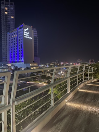 A modern cityscape at night featuring a tall building illuminated with blue and white lights, with the words 'Grand Fiesta Americana' prominently displayed. The foreground shows an elevated walkway with sleek, white railings and embedded ground lights.