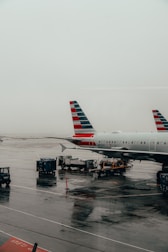A commercial airplane is parked at an airport terminal on a wet tarmac. Ground service vehicles and equipment are positioned near the aircraft for maintenance and loading operations. The plane's tail features a distinct stripe pattern. The atmosphere appears overcast with grey skies.