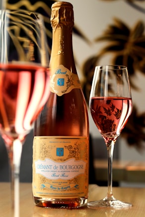 A bottle of Cr&eacute;mant de Bourgogne Brut Ros&eacute; stands prominently on a wooden table, accompanied by two glasses filled with pink sparkling wine. The bottle features a gold label with blue accents, and the reflections of the glasses create a warm and inviting ambiance. In the background, blurred decorative elements add an elegant touch.
