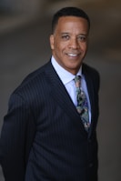 A man is standing confidently, wearing a dark pinstripe suit with a colorful tie. He is smiling slightly and has short, neatly styled black hair. The background appears to be blurred and neutral in color, focusing attention on the person.