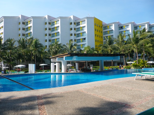 A large swimming pool surrounded by palm trees and lounge chairs with a multi-story hotel building in the background. The building has white walls with yellow accents and multiple balconies. A poolside bar with a canopy and pillars is situated near the pool. The sky is clear and blue, creating a summery and inviting atmosphere.