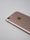 A rose gold smartphone lies on a light gray surface, showing its back side with a camera lens, flash, and the logo in the center. The edges are smooth, and the metallic finish is reflective.