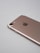 A rose gold smartphone lies on a light gray surface, showing its back side with a camera lens, flash, and the logo in the center. The edges are smooth, and the metallic finish is reflective.