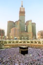 A large crowd of people gathered around the Kaaba, performing a religious ritual in an open courtyard surrounded by arched structures. The Abraj Al Bait Clock Tower rises in the background with its distinctive clock face and towering spire. The scene captures the hustle and devotion of the pilgrims.