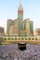 A large crowd of people gathered around the Kaaba, performing a religious ritual in an open courtyard surrounded by arched structures. The Abraj Al Bait Clock Tower rises in the background with its distinctive clock face and towering spire. The scene captures the hustle and devotion of the pilgrims.