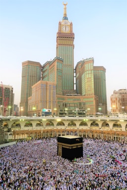 A large crowd of people gathered around the Kaaba, performing a religious ritual in an open courtyard surrounded by arched structures. The Abraj Al Bait Clock Tower rises in the background with its distinctive clock face and towering spire. The scene captures the hustle and devotion of the pilgrims.