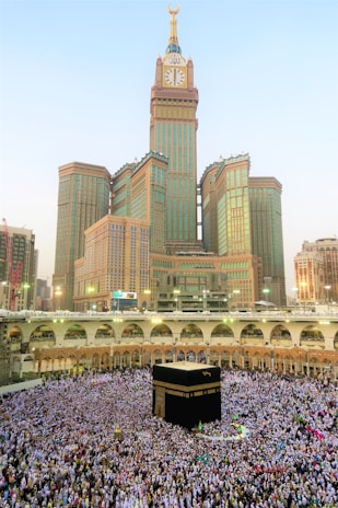 A large crowd of people gathered around the Kaaba, performing a religious ritual in an open courtyard surrounded by arched structures. The Abraj Al Bait Clock Tower rises in the background with its distinctive clock face and towering spire. The scene captures the hustle and devotion of the pilgrims.