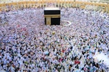 A large crowd of people gathers around the Kaaba within the courtyard of the Masjid al-Haram, engaged in a religious ritual. The people are dressed predominantly in white garments, standing in concentric circles around the central black cube structure, which is adorned with gold calligraphy.