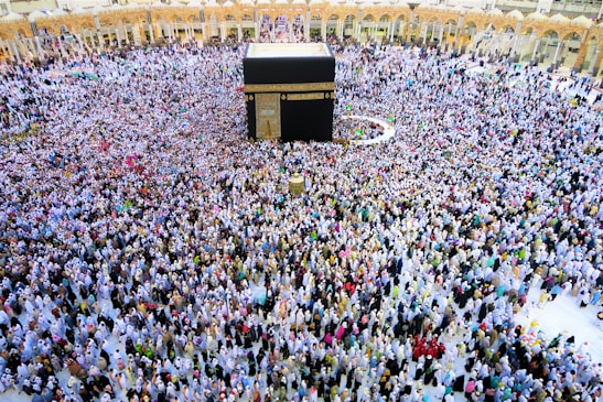 A large crowd of people gathers around the Kaaba within the courtyard of the Masjid al-Haram, engaged in a religious ritual. The people are dressed predominantly in white garments, standing in concentric circles around the central black cube structure, which is adorned with gold calligraphy.