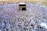 A large crowd of people gathers around the Kaaba within the courtyard of the Masjid al-Haram, engaged in a religious ritual. The people are dressed predominantly in white garments, standing in concentric circles around the central black cube structure, which is adorned with gold calligraphy.