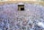 A large crowd of people gathers around the Kaaba within the courtyard of the Masjid al-Haram, engaged in a religious ritual. The people are dressed predominantly in white garments, standing in concentric circles around the central black cube structure, which is adorned with gold calligraphy.