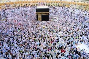 A large crowd of people gathers around the Kaaba within the courtyard of the Masjid al-Haram, engaged in a religious ritual. The people are dressed predominantly in white garments, standing in concentric circles around the central black cube structure, which is adorned with gold calligraphy.