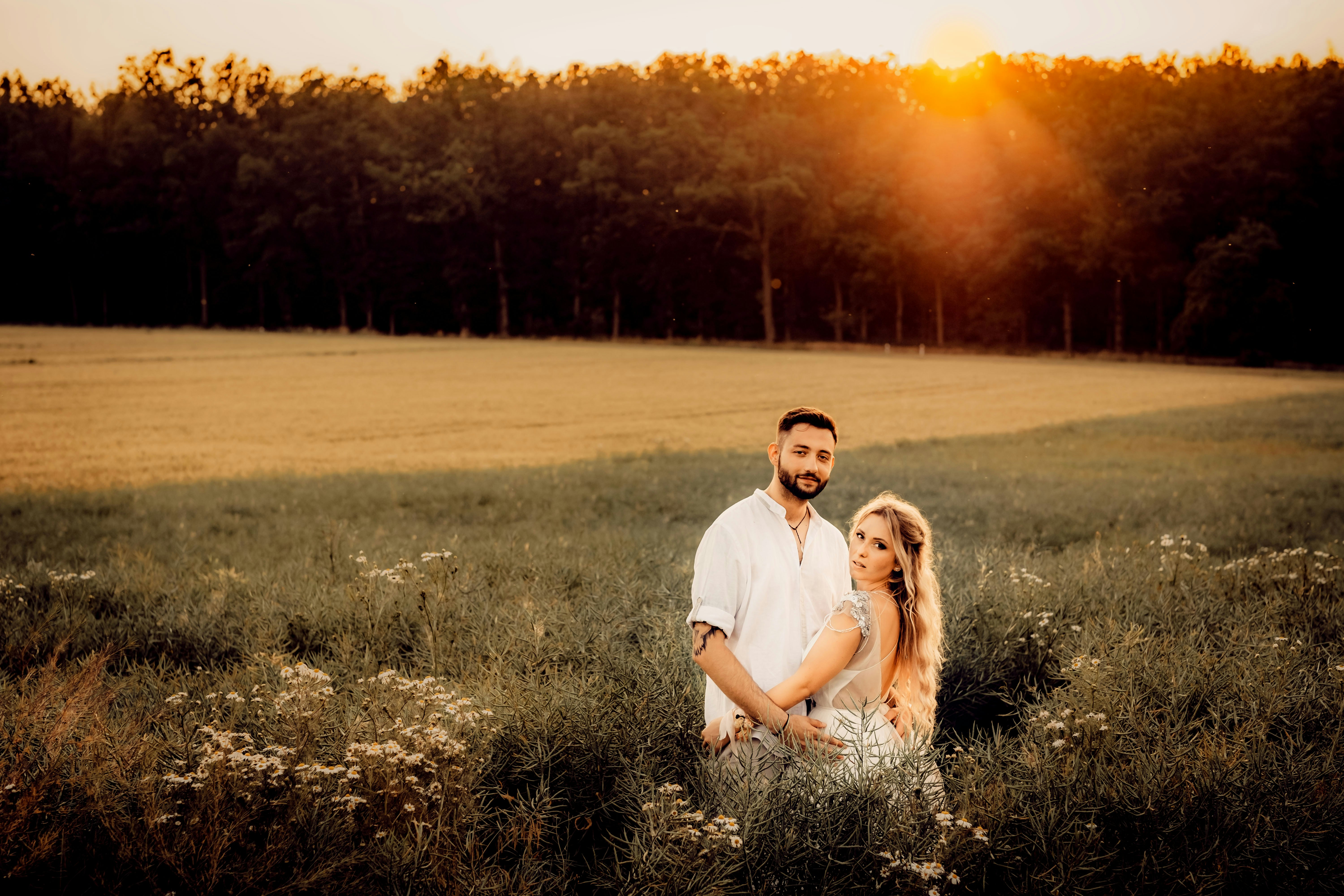 man in white polo shirt hugging woman in white dress on green grass field during sunset