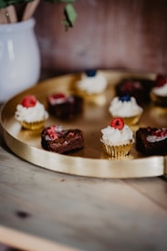 A round golden tray holds several small chocolate cupcakes topped with whipped cream and garnished with fresh raspberries and blueberries. The tray is placed on a wooden surface next to a white ceramic pot containing wooden utensils. The background is softly blurred, highlighting the pastries.
