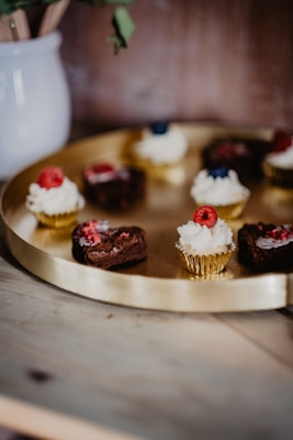 A round golden tray holds several small chocolate cupcakes topped with whipped cream and garnished with fresh raspberries and blueberries. The tray is placed on a wooden surface next to a white ceramic pot containing wooden utensils. The background is softly blurred, highlighting the pastries.