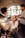 A woman is getting her makeup and hair done by two stylists in a well-lit room with a large window in the background. She is smiling and appears to be enjoying the process. The setting is elegant with a cozy and intimate atmosphere.