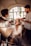 A woman is getting her makeup and hair done by two stylists in a well-lit room with a large window in the background. She is smiling and appears to be enjoying the process. The setting is elegant with a cozy and intimate atmosphere.