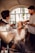 A woman is getting her makeup and hair done by two stylists in a well-lit room with a large window in the background. She is smiling and appears to be enjoying the process. The setting is elegant with a cozy and intimate atmosphere.