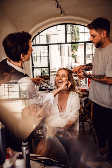 A woman is getting her makeup and hair done by two stylists in a well-lit room with a large window in the background. She is smiling and appears to be enjoying the process. The setting is elegant with a cozy and intimate atmosphere.