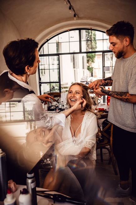 A woman is getting her makeup and hair done by two stylists in a well-lit room with a large window in the background. She is smiling and appears to be enjoying the process. The setting is elegant with a cozy and intimate atmosphere.