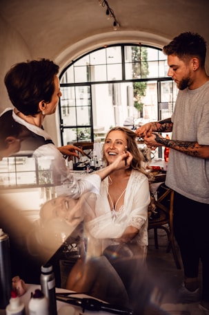 A woman is getting her makeup and hair done by two stylists in a well-lit room with a large window in the background. She is smiling and appears to be enjoying the process. The setting is elegant with a cozy and intimate atmosphere.