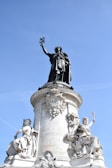 A grand statue of a figure holding a symbol aloft stands atop a stone pedestal with intricate carvings and inscriptions. Two additional sculptures are seated at the base, featuring detailed classical attire and symbols of enlightenment and strength. The backdrop is a clear blue sky, adding contrast to the ornate stonework.