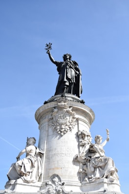 A grand statue of a figure holding a symbol aloft stands atop a stone pedestal with intricate carvings and inscriptions. Two additional sculptures are seated at the base, featuring detailed classical attire and symbols of enlightenment and strength. The backdrop is a clear blue sky, adding contrast to the ornate stonework.