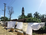 A rustic outdoor scene featuring laundry hung on a line to dry. White sheets and garments are displayed against a backdrop of lush green trees, a tall tower, and a clear blue sky. A cobblestone path runs alongside the drying clothes, with a utility pole and wires visible on the left.