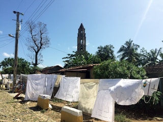 A rustic outdoor scene featuring laundry hung on a line to dry. White sheets and garments are displayed against a backdrop of lush green trees, a tall tower, and a clear blue sky. A cobblestone path runs alongside the drying clothes, with a utility pole and wires visible on the left.