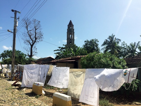 A rustic outdoor scene featuring laundry hung on a line to dry. White sheets and garments are displayed against a backdrop of lush green trees, a tall tower, and a clear blue sky. A cobblestone path runs alongside the drying clothes, with a utility pole and wires visible on the left.