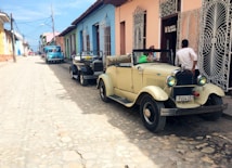 A cobblestone street lined with colorful colonial-style buildings. Two vintage cars, one cream and one black, are parked along the side of the road, serving as taxis. A few people are near the cars, and an old blue truck is visible further down the street. The sky is clear, adding to the bright, nostalgic atmosphere.
