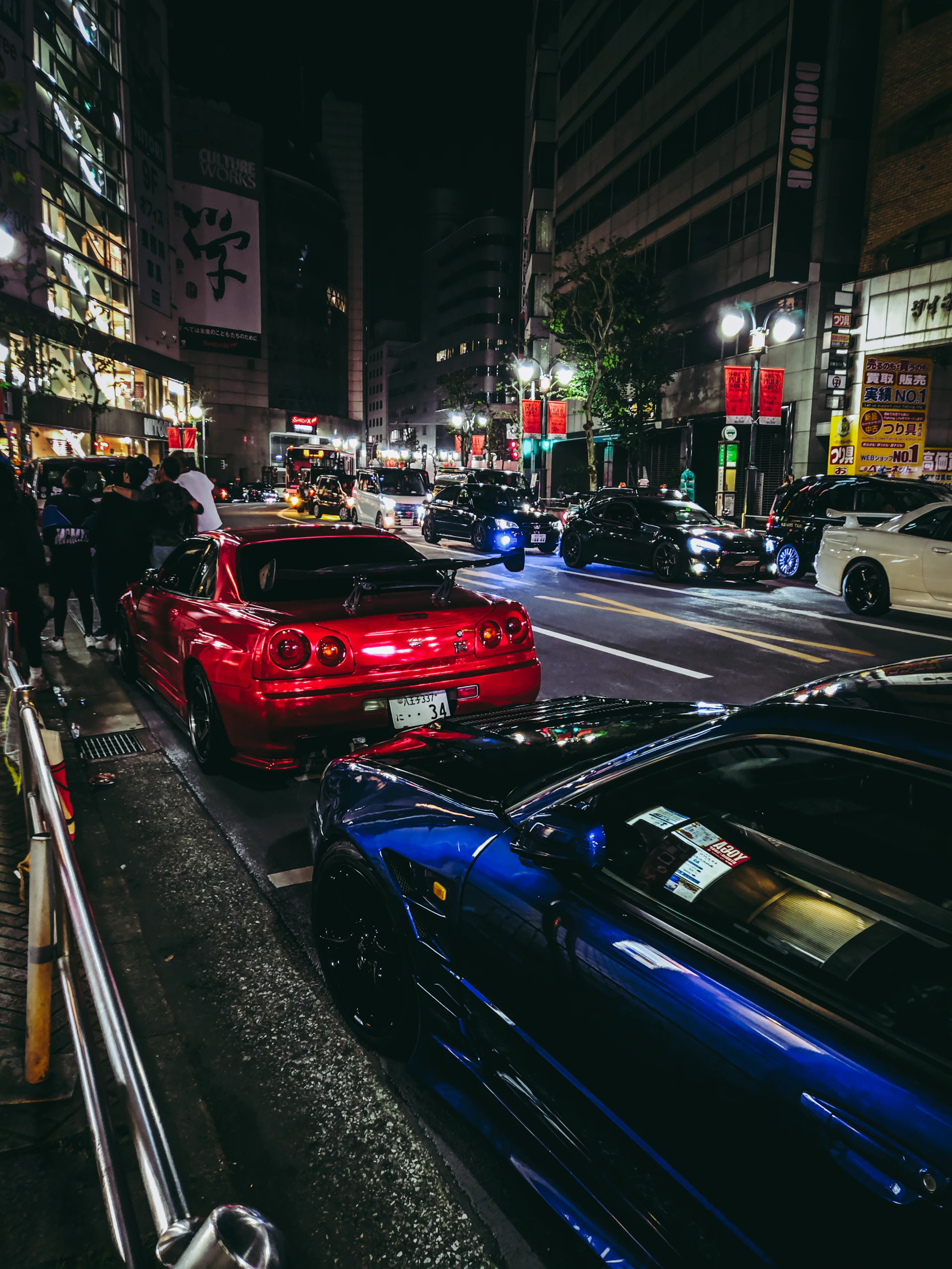 A red and a blue sports car parked on a busy Tokyo street at night