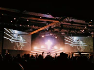 A large crowd of people in silhouette are gathered in front of a stage with a bright cross-shaped light. Two large screens on either side display lyrics, hinting at a musical or worship event. The venue appears to be a concert hall with stage lighting overhead creating an energetic atmosphere.