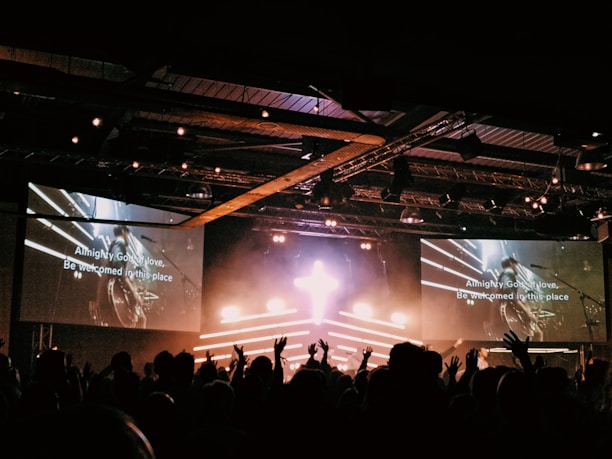A large crowd of people in silhouette are gathered in front of a stage with a bright cross-shaped light. Two large screens on either side display lyrics, hinting at a musical or worship event. The venue appears to be a concert hall with stage lighting overhead creating an energetic atmosphere.