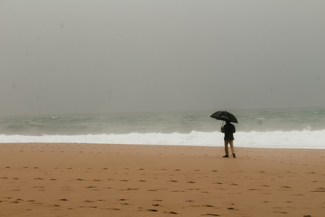 person in black jacket holding umbrella walking on beach during daytime