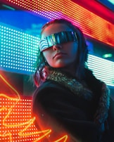 A person wearing futuristic, reflective sunglasses stands against a vibrant background of colorful LED lights. The lights create a neon glow with red, blue, and green hues. The person’s attire includes a coat with a fur collar, enhancing the modern, cyberpunk atmosphere.