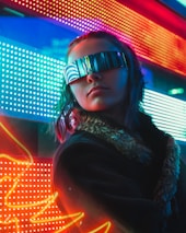 A person wearing futuristic, reflective sunglasses stands against a vibrant background of colorful LED lights. The lights create a neon glow with red, blue, and green hues. The person’s attire includes a coat with a fur collar, enhancing the modern, cyberpunk atmosphere.