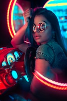 A person wearing reflective sunglasses is seated at an arcade-style racing game console. Surrounding them are vibrant neon lights in red and blue hues, creating a cyberpunk aesthetic. The dashboard of the racing game is detailed with illuminated gauges and controls.