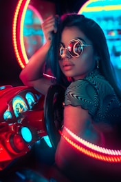 A person wearing reflective sunglasses is seated at an arcade-style racing game console. Surrounding them are vibrant neon lights in red and blue hues, creating a cyberpunk aesthetic. The dashboard of the racing game is detailed with illuminated gauges and controls.