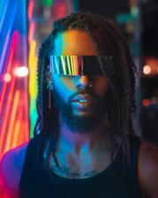 A person with braided hair wearing futuristic, reflective visor glasses, highlighted by vibrant neon lights in the background. The atmosphere feels futuristic and high-tech with glowing colors lighting up the scene.