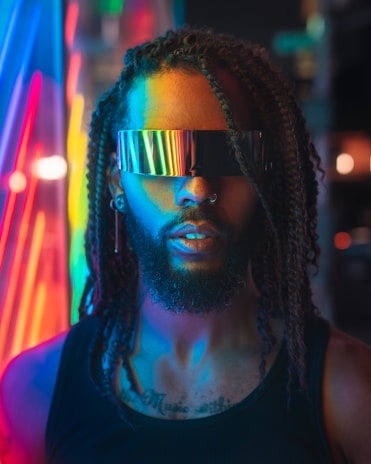 A person with braided hair wearing futuristic, reflective visor glasses, highlighted by vibrant neon lights in the background. The atmosphere feels futuristic and high-tech with glowing colors lighting up the scene.