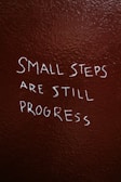 Text is written on a textured, dark brown wall. The phrase reads, 'Small steps are still progress' in white, slightly uneven handwriting.
