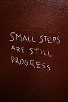 Text is written on a textured, dark brown wall. The phrase reads, 'Small steps are still progress' in white, slightly uneven handwriting.